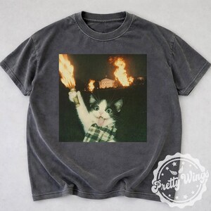 Puede incluir: Camiseta gris oscuro con un gráfico de un gato blanco y negro sosteniendo una antorcha encendida, con un edificio en llamas al fondo. El gato lleva una bufanda a cuadros y saca la lengua.