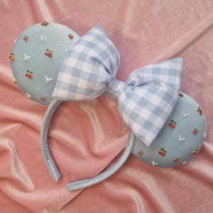 May include: A light blue headband with two round ears and a large bow. The ears are decorated with small red cherries and white pearls. The bow is a blue and white gingham pattern. The headband is on a pink velvet surface.