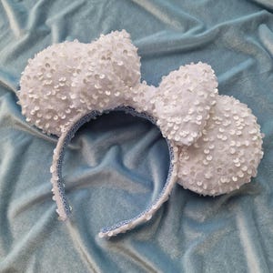 May include: A headband featuring two large, round ears covered in white sequins and a central white sequined bow. The inner band is lined with a light blue fabric featuring a decorative pattern, while the outer edge is trimmed with white sequins.
