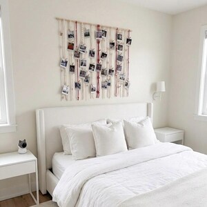 May include: A bedroom with a white bed, pillows, and bedding. A macrame wall hanging with photos is above the bed. A white nightstand with a small device sits to the left of the bed. The room has a neutral color palette.