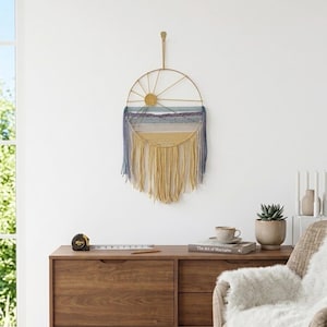 May include: A circular wall hanging with a sun and mountain design. The artwork features a gold metal frame, with blue, yellow, and beige yarn. The piece is hanging on a white wall above a wooden cabinet.
