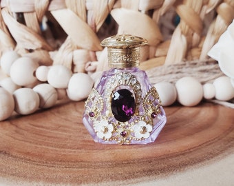 May include: A small, ornate purple glass bottle with a gold lid and a large purple gemstone. The bottle is decorated with gold filigree and white flowers.