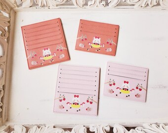 May include: Four card holders with different designs. Each card holder has a pink or orange background with a white animal wearing a red bow and surrounded by red berries and leaves. The card holders have multiple slots for cards.