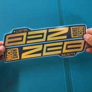 Peut inclure: Un autocollant noir et doré avec le texte "ZEE" et "ROCK SHOX SELECT+". L'autocollant est tenu par des mains sur une surface bleue. L'autocollant a une forme rectangulaire avec des coins arrondis.