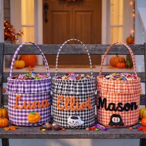 May include: Three fabric buckets with gingham patterns in purple, black, and orange. Each bucket features a ruffled top edge, a handle, and a personalized name in felt lettering: Luna, Elliot, and Mason. Small felt icons of a pumpkin, ghost, and figure are attached.