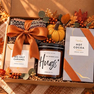 May include: A gift box containing a patterned knit item with a rust-colored ribbon, a yellow knit pumpkin, a candle labeled No Words Just Hugs, a box of Sea Salt Caramel cookies, and a box of Grand Provisions Hot Cocoa with autumn foliage.