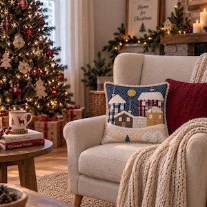 Puede incluir: Un cojín decorativo cuadrado con una escena de pueblo invernal acolchada, casas con patrones en azul, rojo, marrón y amarillo sobre un cielo azul oscuro con puntos y una luna amarilla, acabado con un borde de ganchillo color crema.
