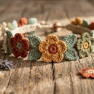 Puede incluir: Una corona floral de ganchillo con una banda color crema adornada con flores de ganchillo coloridas y hojas verdes. Las flores son en tonos naranja, amarillo y verde. La corona se muestra sobre una superficie de madera rústica.