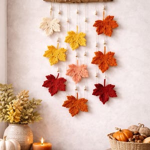 May include: A fall-themed wall hanging featuring crocheted maple leaves in shades of cream, yellow, orange, and red. The leaves are suspended from a wooden branch with beaded accents. A candle, pumpkins, and a vase of foliage complete the autumn decor.