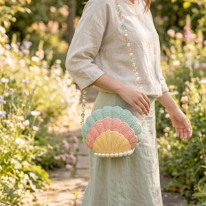 May include: A crochet handbag shaped like a seashell, in pastel shades of yellow, pink, and blue-green. The bag has a pearl beaded trim and strap. The strap is decorated with clear and pastel-coloured beads. The bag is held by a person.