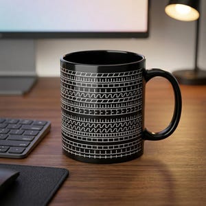 Könnte beinhalten: Ein schwarzer Keramikbecher mit einem sich wiederholenden weißen Grafikmuster aus horizontalen Reifenprofilen und geometrischen Linien. Der Becher hat einen zylindrischen Körper mit einem geschwungenen Henkel auf der rechten Seite.