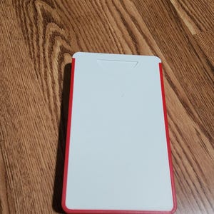 May include: A rectangular white card with a red border, featuring a notch at the top. The card is resting on a wooden surface with a brown wood grain pattern.