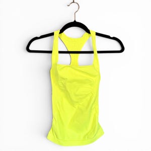 May include: A neon yellow racerback tank top with a square neckline and a perforated detail on the upper back panel. The garment features side ruching and is displayed on a black plastic hanger against a plain white background.