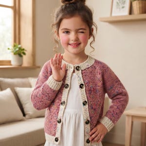 May include: A long-sleeved, mauve-coloured knit cardigan featuring a speckled, multi-coloured pattern throughout. The garment has a cream-coloured ribbed trim along the neckline, cuffs, and hem, and is fastened with six dark, round buttons down the front.