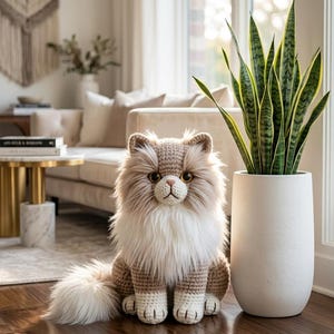 May include: A crocheted plush cat with long, fluffy beige and white fur, amber eyes, and a pink nose. The cat is sitting upright with a full, textured tail. The text DigitalStitchGifts is visible at the top center of the image.