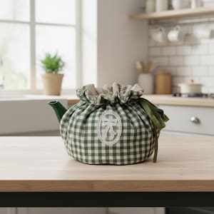 May include: A green and white checkered teapot cozy with a decorative bow design. The cozy is made of fabric and covers a green teapot. A green ribbon is tied to the side. The teapot is on a wooden table in a kitchen setting.