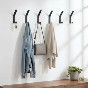 May include: A row of six black metal wall hooks. A denim jacket, a gray purse, and a beige blanket hang from the hooks. A set of keys hangs from the first hook. A small potted plant sits on a wooden surface.