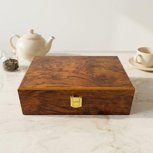 May include: A rectangular wooden box with a dark brown burl wood grain pattern and a polished finish. The box features a gold-coloured metal latch centred on the front edge, designed to keep the lid securely closed for storage and organisation.
