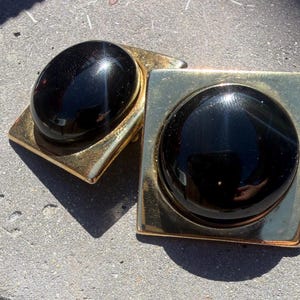 May include: A pair of gold-toned cufflinks with large, oval and round, glossy black stones. The cufflinks have a square base with a gold finish. The stones are set in the center of the square base.