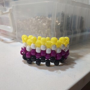 May include: A handmade kandi bracelet featuring yellow, white, purple, and black beads. The bracelet is made with a woven pattern, and the beads are cylindrical. The bracelet is sitting on a white surface.
