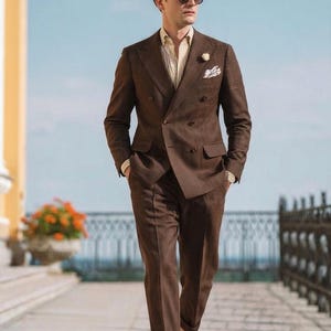 May include: A brown double-breasted suit with a light yellow striped shirt, pocket square, and sunglasses. The suit has a tailored fit and is worn with brown leather loafers. The background features a blue sky and a decorative railing.