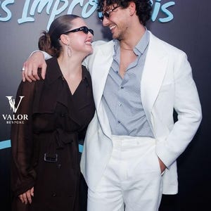 May include: A man in a white suit and sunglasses smiles at a woman in a brown coat and sunglasses. The woman has her hair pulled back. The background is dark with the words "Les Improvis" in light blue.