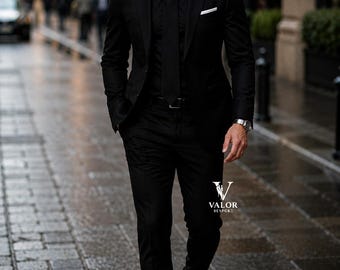 May include: A black suit consisting of a tailored blazer, matching trousers, a black button-down shirt, and a black tie. A white pocket square is tucked into the blazer pocket. The logo VALOR BESPOKE is visible on the lower right side of the suit.