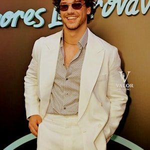 May include: A man in a white suit, a striped shirt, and sunglasses. The suit is a classic design with a two-button jacket and matching trousers. The background has teal text. The man is smiling.