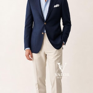 Puede incluir: Un blazer azul marino con botones dorados, combinado con una camisa azul claro y pantalones beige. Un pañuelo de bolsillo blanco añade un toque de elegancia. El conjunto se completa con mocasines de cuero marrón. El logotipo de Valor Bespoke es visible.