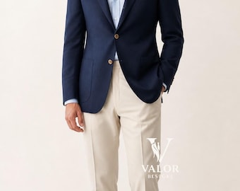 May include: A navy blue blazer with gold buttons, paired with a light blue dress shirt and beige pants. A white pocket square adds a touch of elegance. The outfit is completed with brown leather loafers. The Valor Bespoke logo is visible.