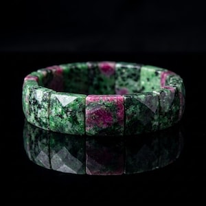May include: A stretch bracelet composed of faceted, rectangular beads made of green zoisite with embedded magenta ruby inclusions. The beads are arranged in a continuous circular band, showcasing a polished, reflective surface with natural mineral patterns.