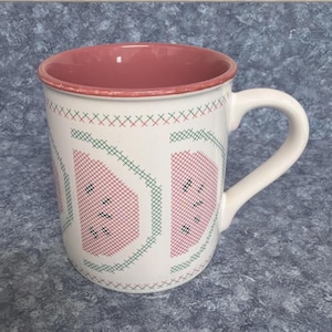 Può includere: Tazza in ceramica bianca con disegno di anguria. La tazza presenta un interno e un manico rosa, con un motivo a fette di anguria in stile punto croce rosso e verde. La tazza è di dimensioni standard per caffè o tè.