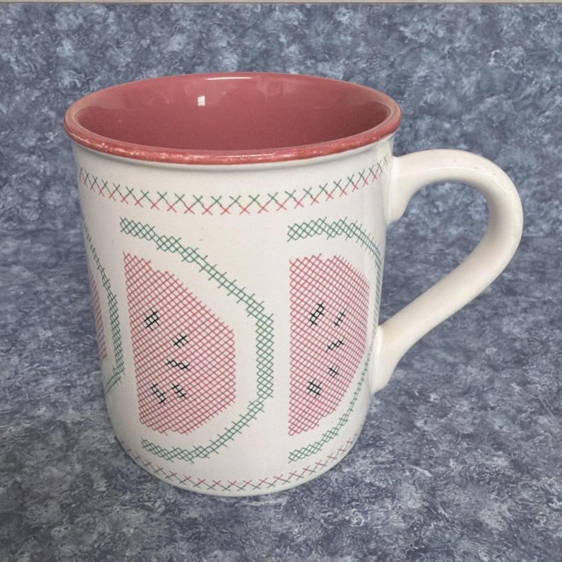 May include: White ceramic mug with a watermelon design. The mug features a pink interior and handle, with a watermelon slice pattern in red and green cross-stitch style. The mug is a standard size for coffee or tea.