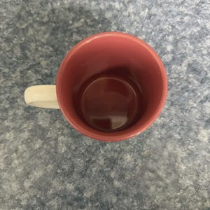 May include: A ceramic mug with a cream-colored handle and a dark pink interior. The exterior of the mug is also dark pink. The mug is empty and sits on a gray speckled surface.
