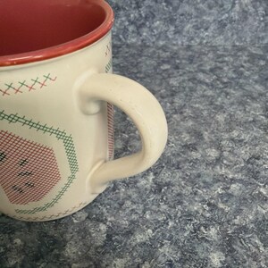 May include: Cream-colored ceramic mug with a red interior and handle. The mug features a cross-stitch design in red and green, with a red rim. The handle is curved and attached to the side of the mug.