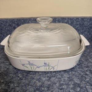May include: White rectangular casserole dish with a clear glass lid. The dish features a floral design with purple and green irises. The lid has a round handle and the dish has white handles on each side.