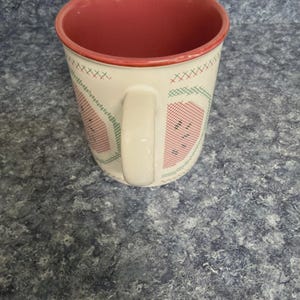 May include: White ceramic mug with a red interior and handle. The exterior features a watermelon design with red, green, and black cross-stitch patterns. The mug is set against a blue and gray speckled surface.