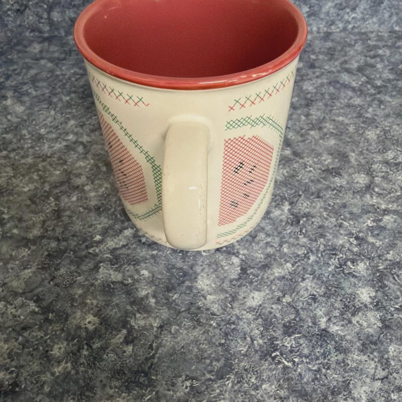 May include: White ceramic mug with a red interior and handle. The exterior features a watermelon design with red, green, and black cross-stitch patterns. The mug is set against a blue and gray speckled surface.