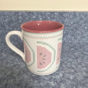 May include: White ceramic mug with a watermelon design. The mug features a red interior and a white handle. The exterior has a cross-stitch pattern with watermelon slices in red and green.