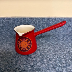 May include: A red coffee pot with a long handle and a white interior. The pot features a sun design with an orange face and yellow rays. The pot is likely made of metal and is designed for stovetop use.