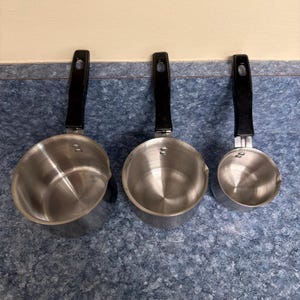 May include: Three silver-colored metal saucepans of varying sizes arranged in a row. Each saucepan features a polished interior, a small pouring spout on the rim, and a long, black, heat-resistant handle with a hole at the end for hanging storage.