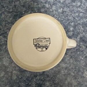 May include: Off-white ceramic mug with a white handle. The bottom of the mug features a black logo with the text "Stitchin' Time Collection" and "Mount Clemens Pottery Made in Japan."