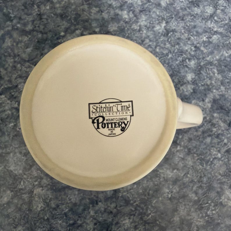 May include: Off-white ceramic mug with a white handle. The bottom of the mug features a black logo with the text "Stitchin' Time Collection" and "Mount Clemens Pottery Made in Japan."