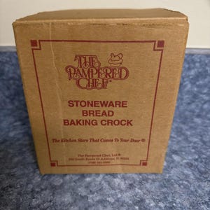 Może przedstawiać: Brązowe kartonowe pudełko na kamionkowy garnek do pieczenia chleba. Pudełko ma czerwoną obwódkę i tekst "The Pampered Chef" oraz "Stoneware Bread Baking Crock". Pudełko zawiera również tekst "The Kitchen Store That Comes To Your Door".