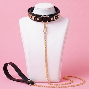 May include: A black collar featuring gold-toned metal lettering that spells out GOOD GIRL, accented with a central gold heart-shaped ring. A matching gold chain leash is attached to the heart ring, ending in a black handle with gold rivets.