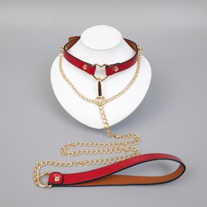Puede incluir: Un collar de cuero rojo con un corazón dorado en el centro y detalles de cadena dorada. El conjunto incluye una correa de cuero roja a juego con una cadena dorada. El collar se exhibe en una cabeza de maniquí blanca.