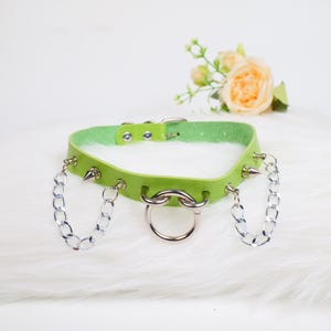 Puede incluir: Un collar gargantilla verde lima con detalles de cadena plateada y un anillo grande. La gargantilla tiene púas plateadas y un cierre de hebilla. El collar se muestra sobre una superficie blanca y esponjosa.