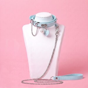 Puede incluir: Un collar de cuero azul claro con herrajes plateados, que incluye una cadena, un dije esférico y un colgante en forma de corazón. Una correa de cadena plateada larga con un asa de cuero azul claro a juego está unida al anillo lateral.