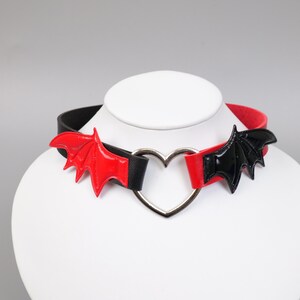 Puede incluir: Un collar gargantilla negro y rojo con detalles de alas de murciélago. El collar tiene un centro en forma de corazón plateado y se muestra en una cabeza de maniquí blanca. Las alas de murciélago son rojas y negras.