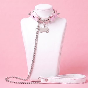 May include: A light pink collar featuring silver-toned metal spikes and a matching silver chain leash. The collar includes a bone-shaped metal tag with a small paw print engraving. The leash has a white textured handle with pink stitching.